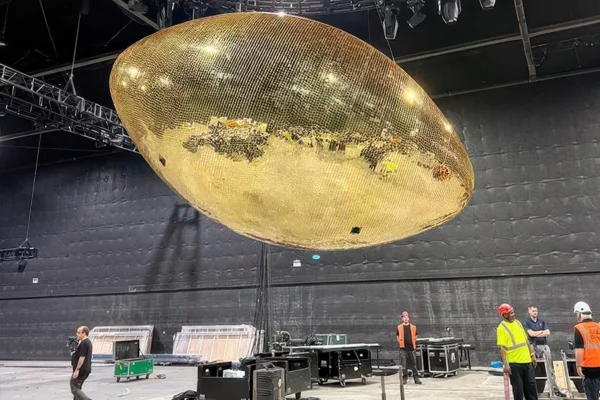 Giant gold mirror football suspended from venue ceiling during load-in